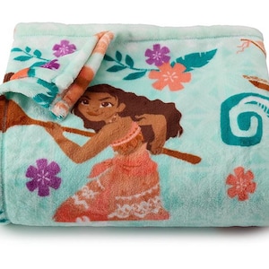 May include: A folded turquoise blanket featuring a cartoon illustration of a woman holding a wooden oar. The blanket has floral and leaf patterns in pink, purple, and teal. The blanket is made of a soft, plush material.