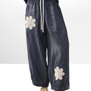 May include: Navy and white striped trousers with a drawstring waist and two cream crochet flower appliqués. The wide-leg trousers have a relaxed fit and are worn with beige sandals.
