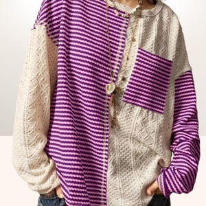 Peut inclure: Un pull à manches longues au design unique. Un côté présente un tissu en tricot ouvert de couleur crème, tandis que l'autre côté et la poche sont rayés de violet et de rose clair. Un collier doré avec un pendentif en forme de cœur est visible.