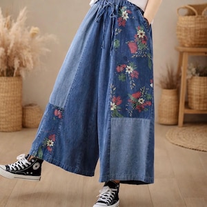 Può includere: Pantaloni ampi in denim blu con ricami floreali. I pantaloni presentano un punto vita con coulisse e un design patchwork. Il ricamo floreale comprende fiori rossi, bianchi e verdi. I pantaloni sono indossati con sneakers alte nere.