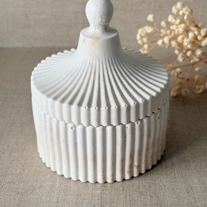 Peut inclure: Un récipient blanc avec un couvercle, présentant un design cannelé et nervuré. La base ronde et le couvercle sont faits d'un matériau solide, avec un petit bouton arrondi sur le dessus. L'ensemble a un aspect légèrement vieilli.