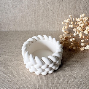 Peut inclure: Un bol décoratif blanc et rond avec un motif de corde tressée texturée sur le bord. Le bol est fait d'un matériau mat et repose sur une surface de couleur neutre. Des fleurs séchées sont en arrière-plan.