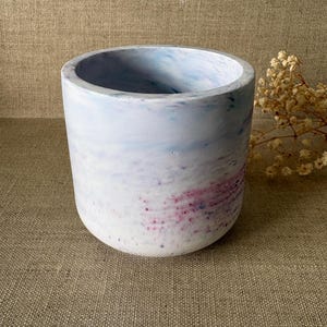 Peut inclure: Un pot de fleurs cylindrique avec un motif marbré. L'extérieur présente un mélange de teintes blanches, bleu clair et roses. Le pot a une base lisse et arrondie et une large ouverture. L'esthétique générale est moderne et minimaliste.