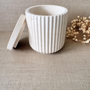 Peut inclure: Un récipient cylindrique blanc avec des détails côtelés verticaux et un couvercle assorti. Le récipient est fait d'un matériau texturé et repose sur une surface de couleur neutre. Des fleurs séchées sont visibles en arrière-plan.
