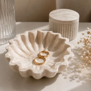 Peut inclure: Un plat en marbre crème à bords festonnés contient deux bagues en or. Un petit pot avec couvercle assorti et un verre texturé sont en arrière-plan. Des fleurs séchées à droite, créant une esthétique neutre et élégante.