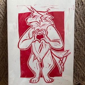 Peut inclure: Une estampe linogravée rouge et blanche représentant une créature anthropomorphe ressemblant à un loup formant un cœur avec ses mains. L'impression est sur papier blanc avec le texte "Puppy Love" et le nom de l'artiste, Sloane.