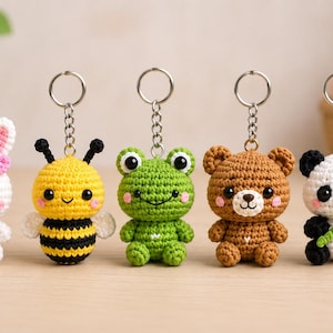 Puede incluir: Cinco llaveros de ganchillo con pequeños animales de peluche: un conejo blanco con lazo rosa, una abeja amarilla y negra, una rana verde, un oso marrón y un panda blanco y negro con un tallo de bambú verde, unidos a anillas de metal plateado.