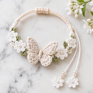 Puede incluir: Una pulsera de ganchillo color crema con un motivo central de mariposa, rodeada de pequeñas flores blancas con centros de perlas y hojas verdes. El cordón ajustable termina en dos flores de ganchillo blancas colgantes con perlas.