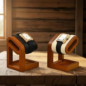 Peut inclure: Deux présentoirs à montres en bois avec des montres. L'une des montres a un bracelet en cuir noir, l'autre un bracelet noir et crème. Les cadrans des montres sont rectangulaires avec des boîtiers dorés. Les présentoirs sont en bois teinté.