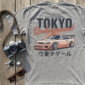 May include: A grey t-shirt featuring a graphic of a modified sports car, the text "TOKYO Underground" with coordinates, and Japanese characters. The design includes a peach-colored car with a rear wing, dark grey lettering, and orange accents.