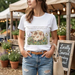 May include: White t-shirt featuring a cartoon cow eating grass, surrounded by flowers. The text on the shirt reads: "There is no ethical consumption under Capitalism (so I am eating grass)." The person is wearing blue jeans and a brown belt.