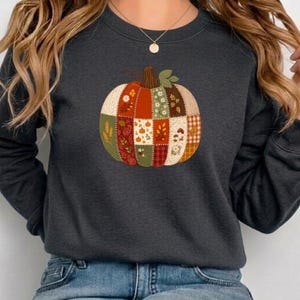 Può includere: Una felpa girocollo grigio antracite scuro con una grande grafica centrale raffigurante una zucca composta da vari motivi in tessuto stile patchwork nelle tonalità dell'arancione, verde, crema e marrone, inclusi motivi floreali, a quadri e spighe.