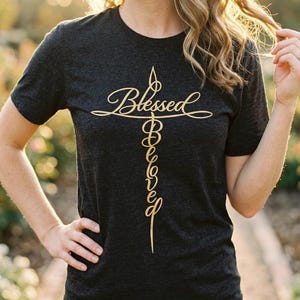 May include: A charcoal grey short-sleeved t-shirt featuring a gold-coloured graphic design of a cross. The word Blessed is written horizontally across the crossbar, while the word Beloved is written vertically down the centre of the cross.