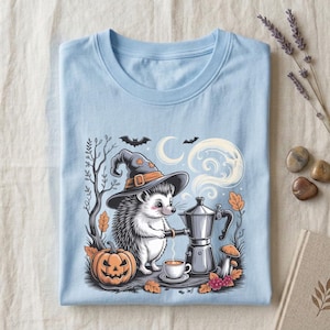 May include: A light blue t-shirt featuring a graphic of a hedgehog wearing a witch hat, pouring coffee from a moka pot into a cup. The scene includes a jack-o'-lantern, a crescent moon, bats, bare trees, mushrooms, and autumn leaves.