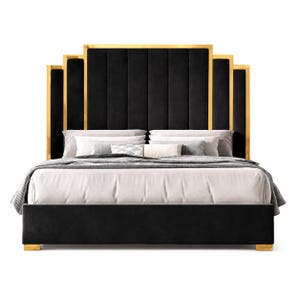 Op de afbeelding: Een zwart gestoffeerd bedframe met een hoog, getrapt hoofdeinde met verticale kanaalstiksels en een goudkleurig metalen frame. Het bed is opgemaakt met wit en grijs beddengoed, inclusief meerdere kussens en een grijze plaid met textuur.