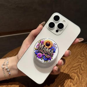 May include: A white smartphone with a floral pop-up grip that says "MAMA". The pop-up grip features a sunflower, blue and purple flowers, and a leopard print design. The phone is held in a hand with a silver bracelet.