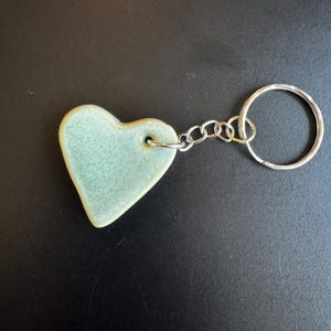 May include: A light green heart-shaped keychain. The heart is made of ceramic and has a glossy finish. It is attached to a silver chain and a large silver keyring. The keychain is on a black surface.