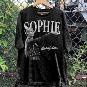 May include: A black short-sleeve t-shirt featuring a distressed white graphic print. The design includes the name SOPHIE in large capital letters above a black and white image of a person pointing, accompanied by a white cursive signature graphic.