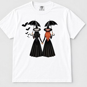 Puede incluir: Una camiseta blanca de manga corta con un gráfico de dos figuras con vestidos negros largos y sombreros puntiagudos, sosteniendo paraguas negros y tomadas de la mano. Una bandada de murciélagos negros vuela hacia la izquierda. Etiqueta Comfort Colors.