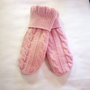 May include: A pair of light pink cable-knit mittens. The mittens have a ribbed cuff and a classic cable-knit design on the front. The soft, warm mittens are perfect for cold weather.