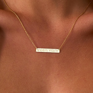 May include: A gold-colored necklace with a rectangular pendant. The pendant has engraved text: "40°42'45.9"N 74°00'21.0"W". The necklace is worn on a person wearing a cream-colored tank top.