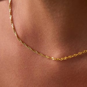 May include: A thin, gold-colored chain necklace featuring a twisted rope-style link design. The necklace rests against the skin, showcasing a metallic yellow finish with a reflective, textured surface that catches the light along its length.