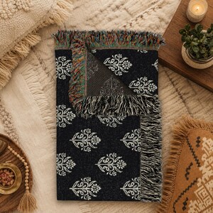 May include: A rectangular woven throw blanket featuring a dark black base with a repeating white floral paisley pattern. The blanket has a folded corner revealing a multicolored fringe edge and a textured reverse side with a lighter color scheme.