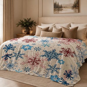 May include: A cream-coloured fleece blanket featuring a repeating pattern of large snowflakes in shades of dark blue, light blue, and deep red. The blanket is draped over a bed, covering the mattress and hanging down over the sides and foot.