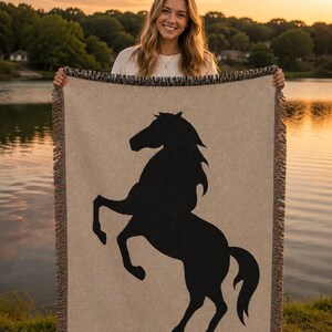 Op de afbeelding: Een rechthoekige geweven plaid met een grote zwarte silhouet van een steigerend paard in het midden op een lichtbeige achtergrond. De deken heeft een geweven textuur en is afgewerkt met decoratieve franjes aan de boven- en onderkant.