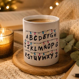 Op de afbeelding: Witte keramische mok met zwart alfabet en kleurrijke lichtslingers. De mok, gevuld met koffie, staat op een houten onderzetter. Een brandende kaars en een boek zijn op de achtergrond.
