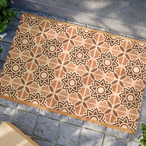 May include: Rectangular doormat with a repeating floral pattern in shades of brown and tan. The design features stylized flower shapes and geometric elements. The mat has a tan border and is placed on a stone surface.