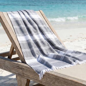 Può includere: Un telo mare a righe grigie e bianche con frange, drappeggiato su una sedia a sdraio di legno. L'asciugamano presenta ampie strisce grigie e bianche alternate con sottili strisce bianche di accento. Lo sfondo mostra una spiaggia sabbiosa e l'oceano.