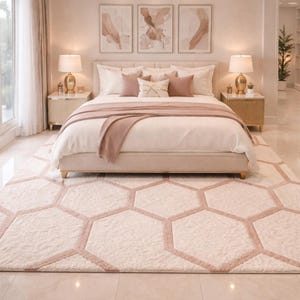 May include: A bedroom with a bed featuring a cream headboard, pillows, and bedding. A blush-coloured throw is draped across the bed. A large rug with a honeycomb pattern in cream and blush tones covers the floor. Two bedside tables with lamps flank the bed.