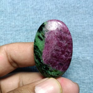 Puede incluir: Una piedra preciosa en forma de cabujón ovalado con una superficie pulida y una clara división de colores. Un lado muestra un tono magenta rosado intenso, mientras que el otro lado presenta un color verde bosque oscuro con inclusiones negras.