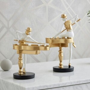 May include: Two decorative figurines of figures playing billiards. The figures are white with gold accents, including hats and the billiard tables. The tables are supported by gold legs and black bases. The background is a light gray and white geometric pattern.