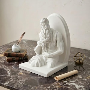 Puede incluir: Un sujetalibros de resina blanca que representa una figura sentada con una larga barba, sosteniendo un libro. El sujetalibros está sobre una superficie de mármol, con un pequeño tintero de mármol y libros a la izquierda. También hay un rollo enrollado y un objeto geométrico.