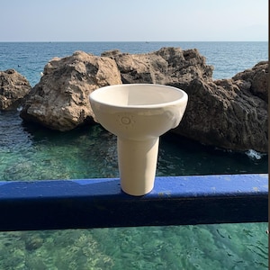 May include: A cream-colored ceramic vessel with a wide, circular opening and a tapered base. The exterior surface features a small, embossed sun icon on the side, and the material has a smooth, matte finish throughout the entire structure.