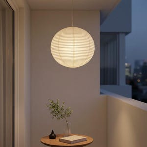 May include: A spherical paper lantern, white in colour, suspended from the ceiling. The lantern's surface is textured with horizontal lines. The scene is set on a balcony, with a small table and a view of a city skyline in the background.