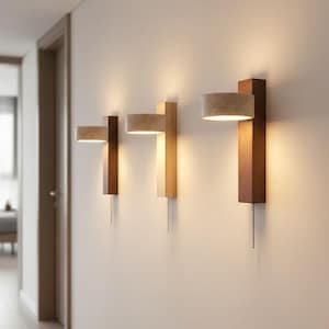 May include: Three wall sconces with a minimalist design. Each sconce features a rectangular wooden base in varying shades of brown and tan, topped with a cylindrical lampshade. A pull chain hangs from each light. The sconces are mounted on a white wall.