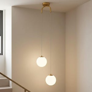 Puede incluir: Dos lámparas colgantes modernas con pantallas esféricas de vidrio blanco. Las luces cuelgan de un accesorio dorado fijado al techo. Las luces están suspendidas por finos cordones, creando una estética minimalista.
