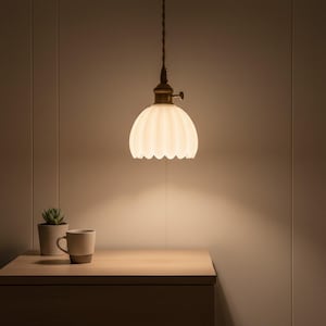 Peut inclure: Une suspension luminaire avec un abat-jour en verre blanc cassé festonné est suspendue au-dessus d'une table de chevet en bois. Le luminaire a une base en métal couleur laiton et un cordon recouvert de tissu. Une petite plante en pot et une tasse sont posées sur la table de chevet.