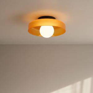 May include: A modern ceiling light fixture with a round, orange-yellow shade and a black base. The light has a white, spherical bulb in the center. The fixture is mounted on a white ceiling.