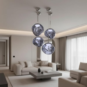 May include: Four modern pendant lights with smoky gray glass globes hang from the ceiling. The globes have a unique, organic shape and are suspended by thin, silver-colored rods. The lights are in a living room with neutral-toned furniture.