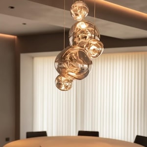 Può includere: Un lampadario moderno con più globi di vetro ambrato di forma irregolare sospesi dal soffitto. I globi variano di dimensioni e riflettono la luce, creando un bagliore caldo. L'apparecchio è sopra un tavolo rotondo.