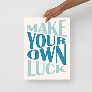Könnte beinhalten: Ein rechteckiger Kunstdruck mit dem Schriftzug "MAKE YOUR OWN LUCK" in Blautönen auf cremefarbenem Hintergrund. Der Druck wird von einer Hand gehalten und zeigt die motivierende Botschaft in einer modernen Schriftart.