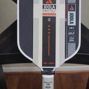 May include: A Joola Pro Perseus pickleball paddle with a black and white striped face. The paddle has the text "JOOLA PRO IV PERSEUS" and "16 MM". The handle has the text "REMOVE FOR TAP HERE".