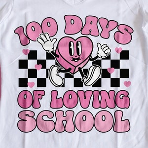 May include: White V-neck t-shirt with a graphic that says "100 Days of Loving School." The graphic features a pink heart character with arms and legs, set against a black and white checkered background with pink hearts.