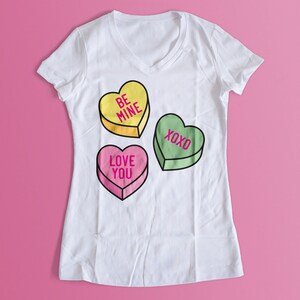 Pu&ograve; includere: T-shirt bianco con scollo a V con tre caramelle a forma di cuore. La caramella gialla dice "BE MINE", la caramella rosa dice "LOVE YOU" e la caramella verde dice "XOXO". La maglietta &egrave; su sfondo rosa.