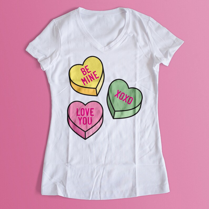 Pu&ograve; includere: T-shirt bianco con scollo a V con tre caramelle a forma di cuore. La caramella gialla dice "BE MINE", la caramella rosa dice "LOVE YOU" e la caramella verde dice "XOXO". La maglietta &egrave; su sfondo rosa.