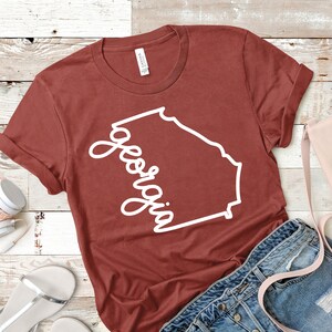 May include: A rust-colored t-shirt with a white outline of the state of Georgia and the word "Georgia" written in a cursive font.